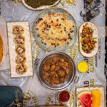 Overhead view of an Afghan meal for two on a plastic-covered carpet: pulao, charsi karahi, mantu, palak, naan, salad and fruit