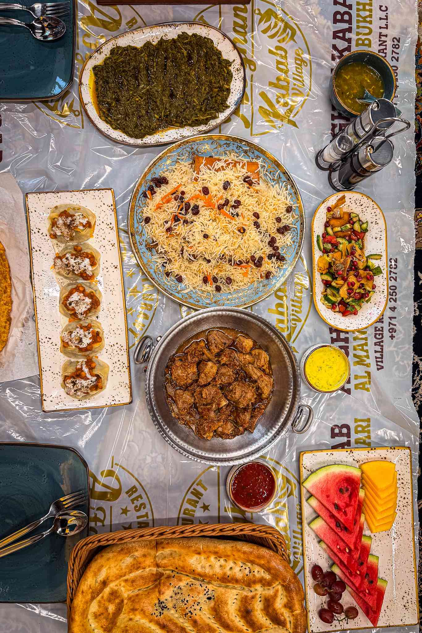 Overhead view of an Afghan meal for two on a plastic-covered carpet: pulao, charsi karahi, mantu, palak, naan, salad and fruit