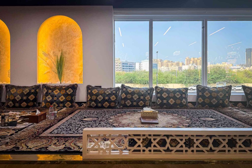 Empty raised carpeted seating platform with bolster cushions and lit arched alcoves beside floor-to-ceiling windows overlooking Deira