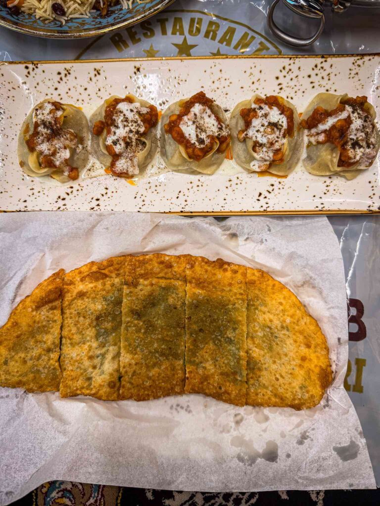 Five Afghan mantu dumplings topped with tomato lentil sauce, yoghurt and dried mint, beside a fried onion bolani cut into fingers