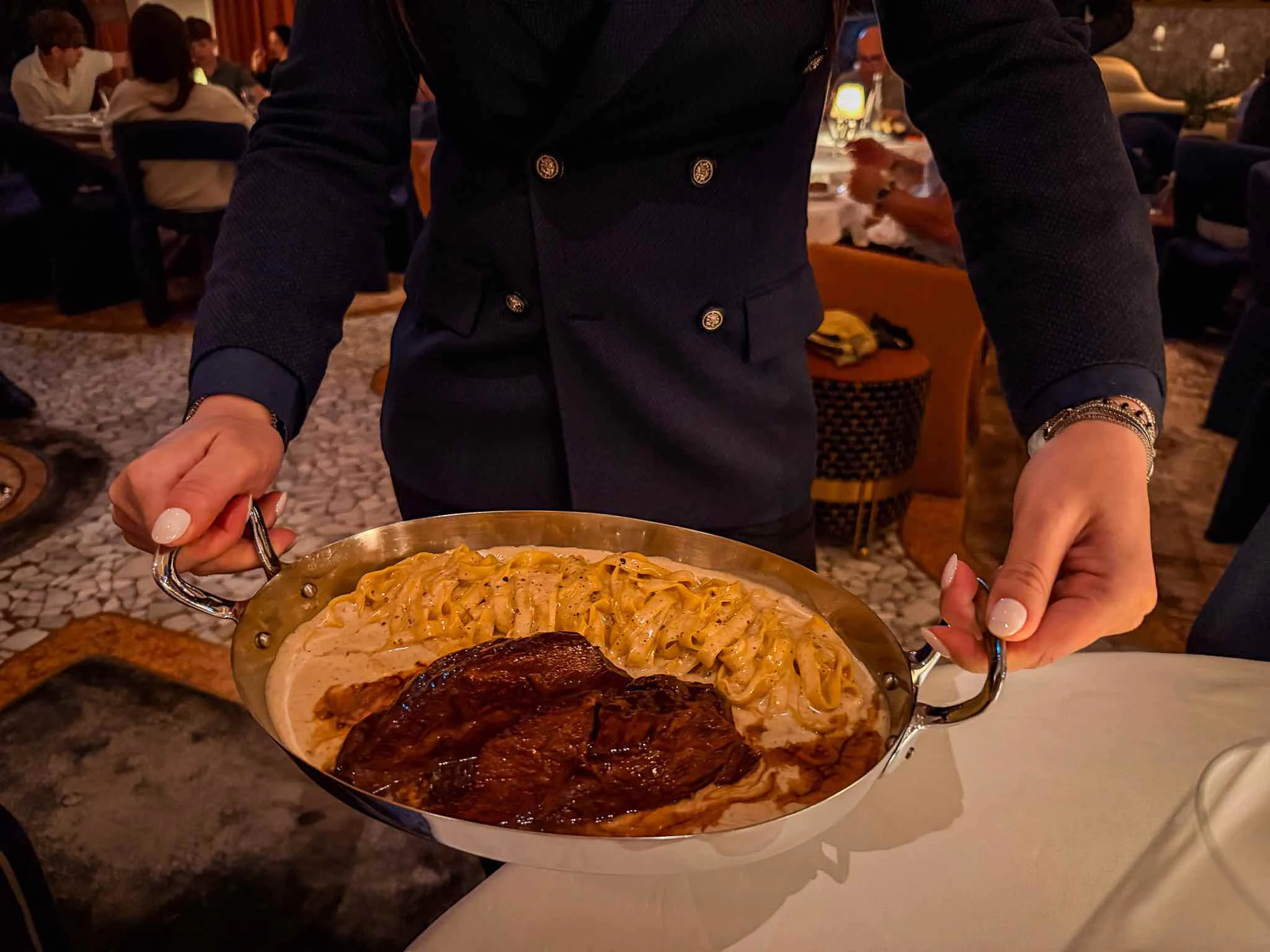 Server holding a large metal pan containing beef cheek pasta and sauce before serving it at the table.