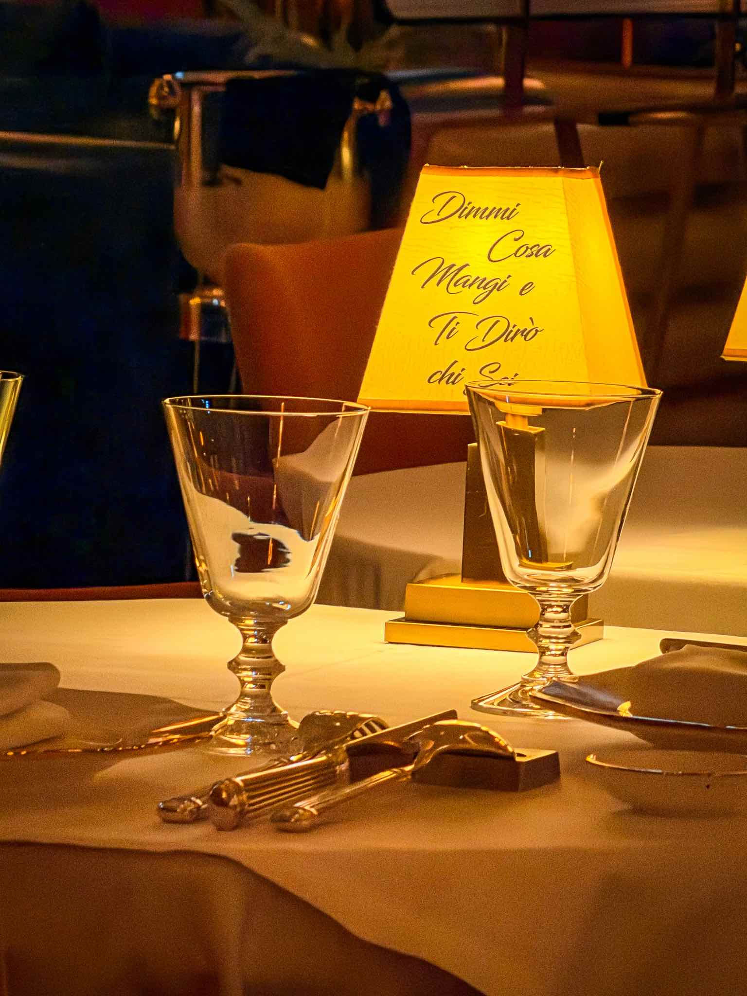 Illuminated table lamp between two glasses on a white-clothed table inside Rialto restaurant in Dubai.