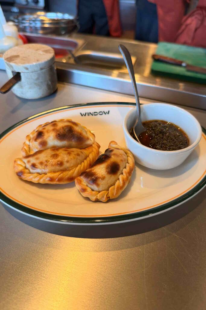 Three baked beef empanadas with braided edges and a pot of chimichurri on a Window-branded plate