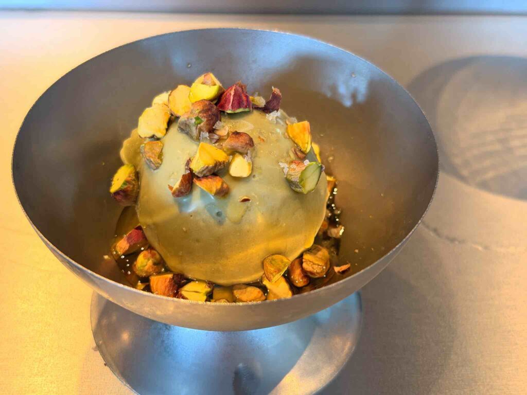 Scoop of pistachio ice cream topped with chopped pistachios, sea salt flakes and olive oil in a footed steel coupe
