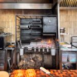 Wood-fired grill with patties cooking over embers, toasted burger buns on a chequered end-grain board in the foreground