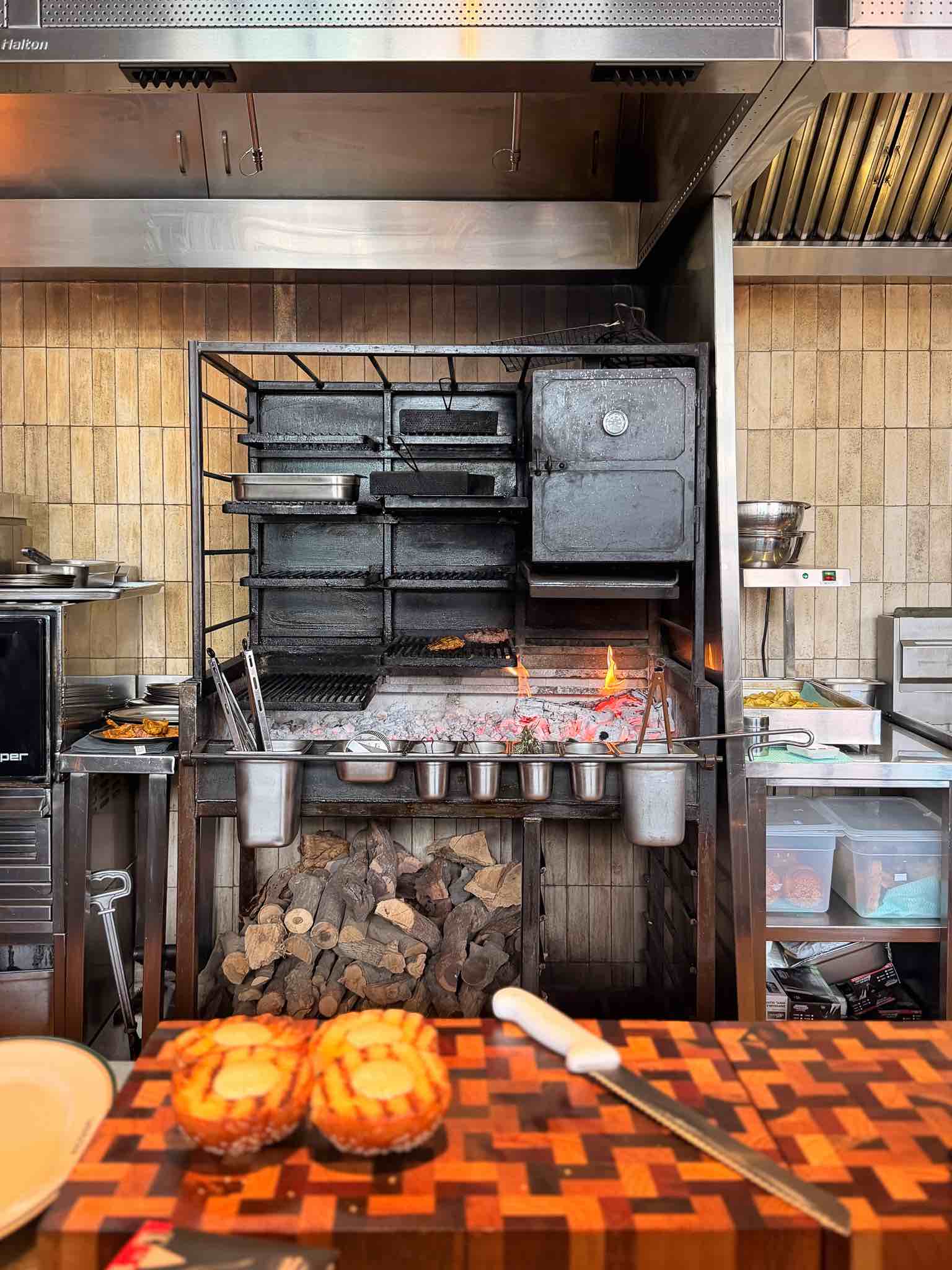 Wood-fired grill with patties cooking over embers, toasted burger buns on a chequered end-grain board in the foreground