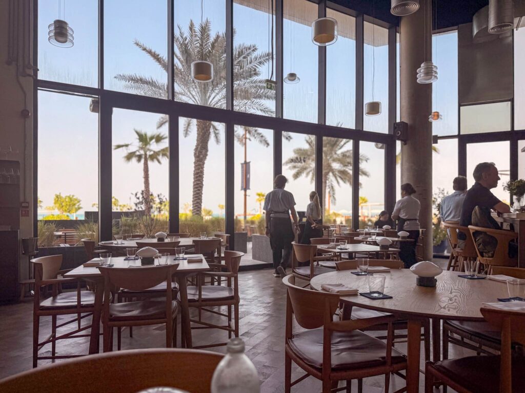 Dining room at Blue restaurant, Mamsha Al Saadiyat, Abu Dhabi, with oak tables and floor-to-ceiling windows onto palms and the sea