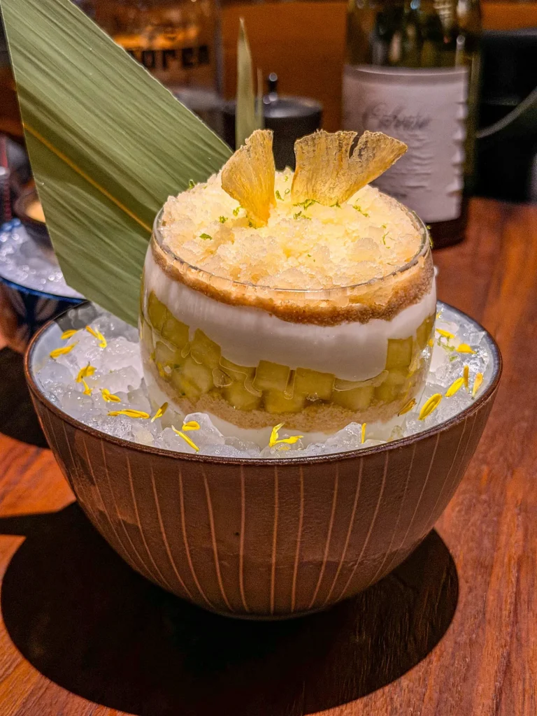 Layered pineapple and chilli dessert at Noren in Dubai: crumb, diced pineapple and cream topped with pineapple granita, lime zest and dried pineapple crisps, on crushed ice with yellow petals.
