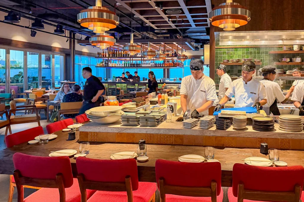Chefs in bandanas working at Noren’s open kitchen in JLT, one waving to the camera, behind a stone counter under copper pendant lamps and green tiled walls.