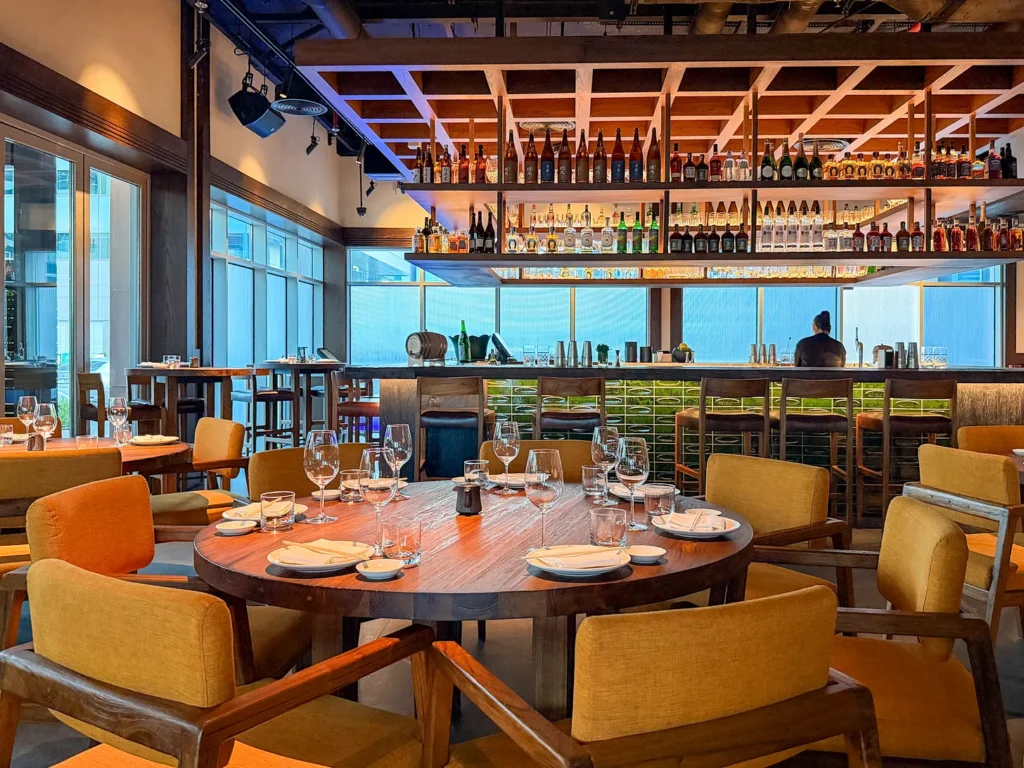 Round table set for dinner in Noren’s dining room in JLT, Dubai, surrounded by mustard armchairs, with a bar of sake and spirits on open shelves against floor-to-ceiling windows