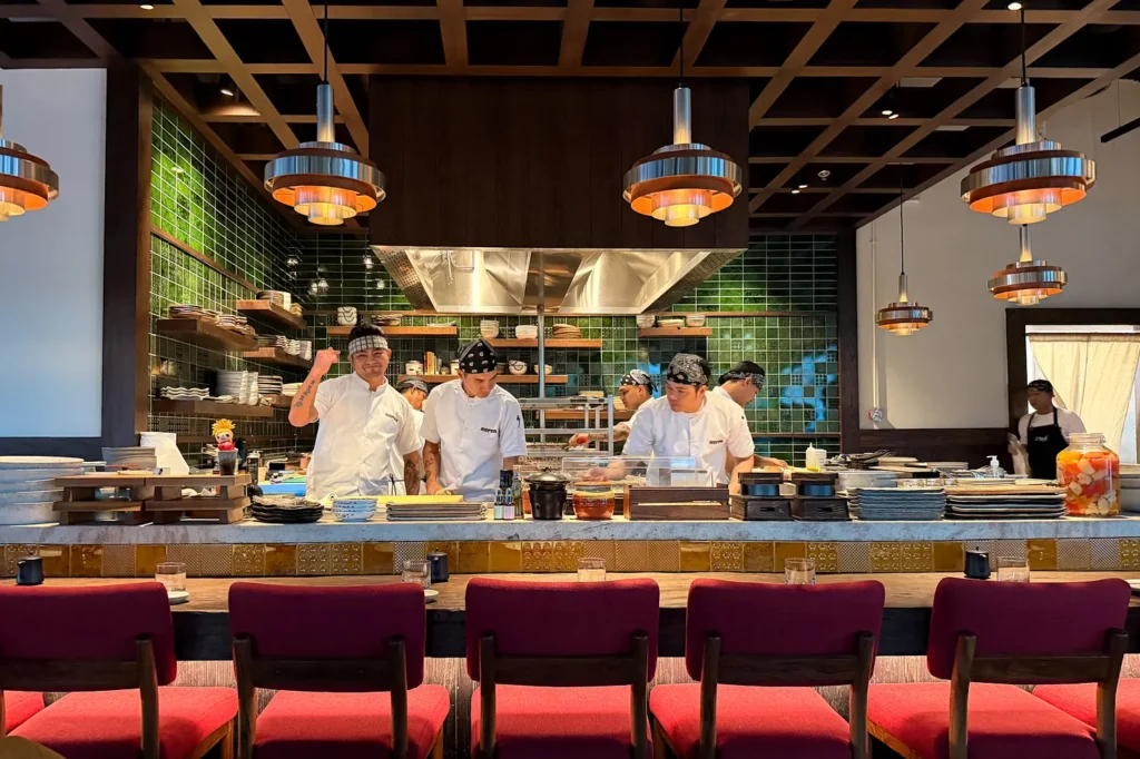 Chefs preparing sushi at the live counter of Noren, a Japanese restaurant in JLT, Dubai, with red counter stools in front, diners at tables and a backlit cocktail bar at the back of the room