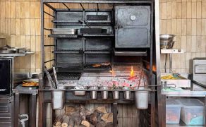 Wood-fired grill with patties cooking over embers, toasted burger buns on a chequered end-grain board in the foreground